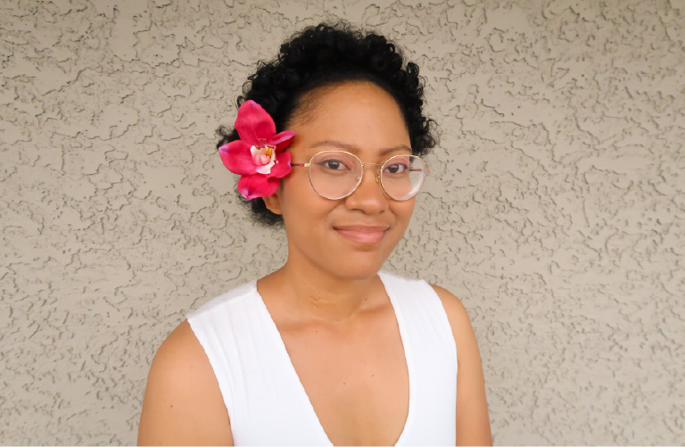 a portrait of the author with a flower tucked behind her right ear. She's wearing a white shirt and thin round glasses. 
            Brown skin and short curly hair.